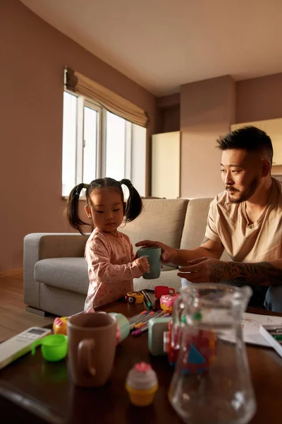 Father taking cup from daughter hands during they playing at table at home. Asian man and girl spend time together. Domestic entertainment and leisure. Family relationship. Parenting and fatherhood