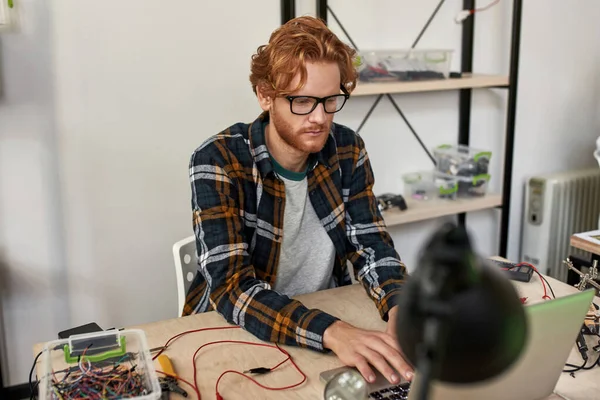 Concentrated smart male IT engineer using laptop in office. Young red haired european man wearing glassed sitting at table with variety technical tools and components. Modern technology and innovation