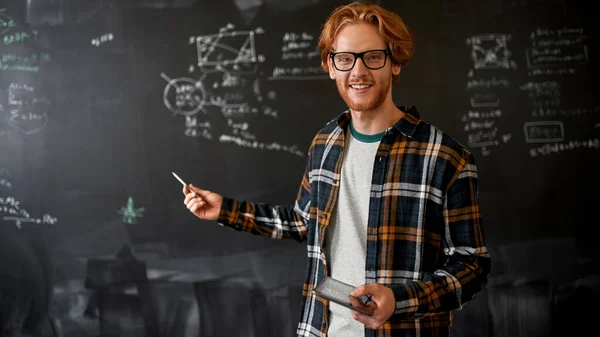 Smiling male teacher pointing with pencil at blackboard with programming codes and formulas in classroom. Young caucasian man with digital tablet wear glasses. IT, technical and engineering education