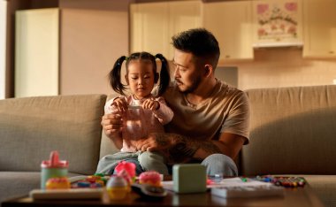 Baba, evdeki kanepedeki dijital termometrede sıcaklığı ölçen odaklanmış kızına bakıyor. Asyalı bir adam ve küçük bir kız. Sağlık ve tıbbi bakım. Aile ilişkisi. Ebeveynlik ve babalık