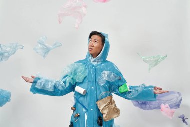 Yağmurluğun altında duran Asyalı hoşnutsuz bir adam. Ekoloji güvenliği ve korunma. Çöp öğütücü ve geri dönüşüm. Çevresel sürdürülebilirlik. Beyaz arka plan. Stüdyo çekimi. Boşluğu kopyala