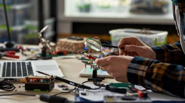 Kırpılmış erkek mühendis mikroçip lehimleyen lehim demiri büyüteç altında. Masada dizüstü bilgisayarla, teknik aletlerle ve parçalarla çalışan bir adam. Modern teknoloji ve yenilik