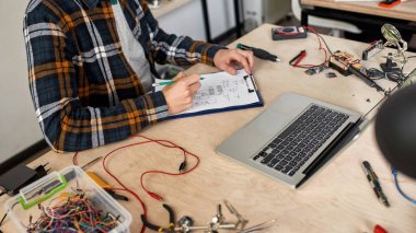 Kısmi erkek bilişim teknisyeni veya mühendis kalem ile teknik düzenleme yapıyor. Ofiste çeşitli araç gereçlerle çalışan bir adam. Modern teknoloji ve yenilik gelişimi