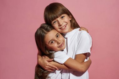 Gülümseyen beyaz küçük kızlar sarılıp kameraya bakıyor. Zoomer kuşağının kız ya da kız kardeşleri. Modern genç yaşam tarzı. Arkadaşlık. Pembe arka planda izole edilmiş. Stüdyo çekimi. Boşluğu kopyala