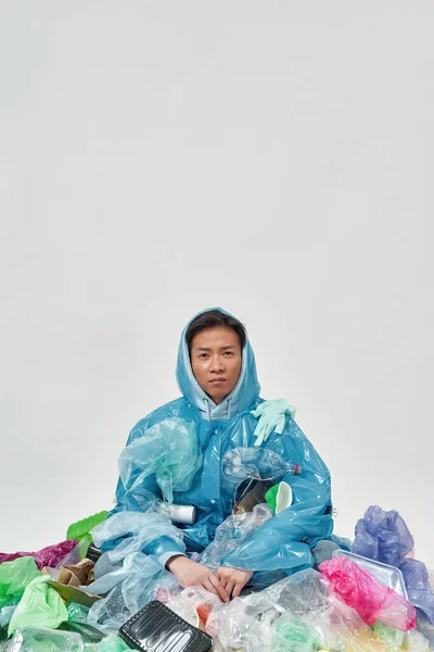 Young asian man wearing raincoat sitting in variety garbage. Ecology safety and protection. Waste disposal and recycling. Environmental sustainability. White background. Studio shoot. Copy space