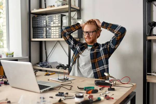 Thoughtful male IT technician or engineer watching laptop in office. Young red haired caucasian man sitting at table with variety technical tools and components. Modern technology and innovation