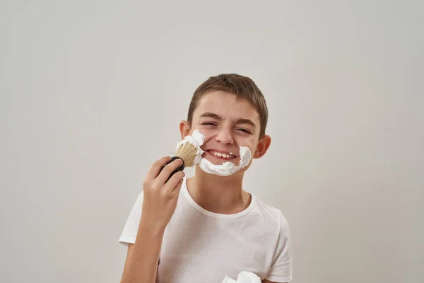 Cropped of smiling caucasian little boy smearing shaving foam on face with brush. Male child of zoomer generation. Modern youngster lifestyle. Skin care. White background. Studio shoot. Copy space