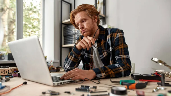Tired male IT technician or engineer using and watching laptop in office. Young red haired caucasian man sitting at table with variety technical tools and components. Modern technology and innovation
