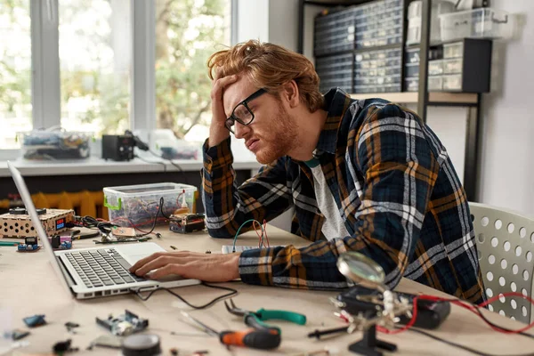 Exhausted male IT technician watching laptop computer in office. Young red haired caucasian man sitting at table with variety technical tools and components. Modern technology and innovation