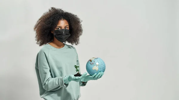 Black girl wearing medical mask and gloves holding earth globe and green plant in ground. Global ecology safety and protection. Waste disposal and recycling. White background. Studio shoot. Copy space