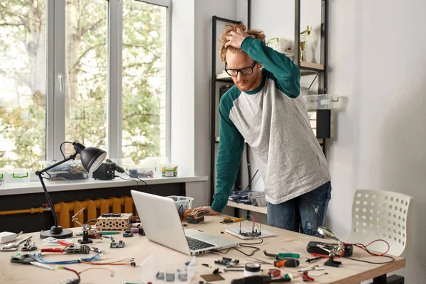 Male IT technician or engineer watching laptop connecting with microchip in office. Young caucasian man standing at table with variety technical tools and components. Modern technology and innovation