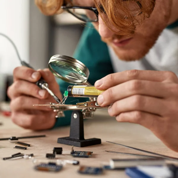 Partial male engineer wearing glasses soldering microchip with soldering iron under magnifying glass at table with blurred variety technical tools and components. Modern technology and innovation