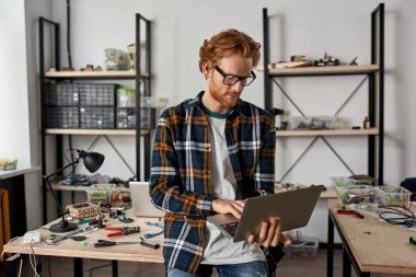 Ofiste dizüstü bilgisayar kullanan konsantre erkek bilişim teknisyeni veya mühendisi. Kafkasyalı gözlüklü genç adam çeşitli teknik aletler ve parçalarla masanın yanında duruyor. Modern teknoloji ve yenilik