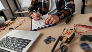 Kesilmiş erkek bilişim mühendisi, kağıt üzerinde kalemle teknik düzenleme yapıyor. Masada dizüstü bilgisayarla, çeşitli teknik aletlerle ve ekipmanlarla çalışan bir adam. Modern teknoloji ve yenilik