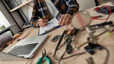 Kısmi erkek bilişim mühendisi kalem ile teknik düzenleme yapıyor. Masada dizüstü bilgisayarla, çeşitli teknik aletlerle ve ekipmanlarla çalışan bir adam. Modern teknoloji ve yenilik gelişimi