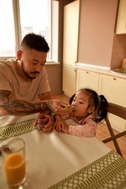 Asyalı baba, küçük kızına ev mutfağında kaşıkla bebek maması veriyor. Aile ilişkisi ve şefkat. Ebeveynlik ve babalık. Sağlıklı beslenme. Ev hayatı işte. Geniş daire