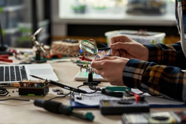 Kısmi erkek mühendis mikroçip lehimlerken lehim demiri büyüteç altında. Masada dizüstü bilgisayarla, teknik aletlerle ve parçalarla çalışan bir adam. Modern teknoloji ve yenilik