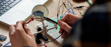 Kısmi erkek bilişim teknisyeni ya da mikroçip lehimleyen lehim aletinin seçici odak noktası büyüteç altında. Masada çalışan bir adam. Modern teknoloji ve yenilik