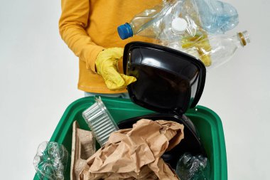 Çöp tenekesinin üzerinde plastik çöp tutan eldivenli bir kız. Çöp ayıklama. Çöp öğütücü ve geri dönüşüm. Çevresel sürdürülebilirlik. Beyaz arka planda izole edilmiş. Stüdyo çekimi