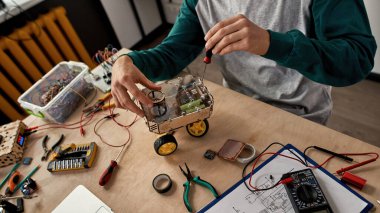 Kısmi erkek bilişim teknisyeni veya mühendis, çeşitli teknik aletler ve parçalarla birlikte masada tornavidayla otomatik tekerlek robotunu tamir ediyor. Modern teknoloji ve yenilik