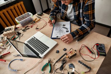 Kesilmiş erkek bilişim teknisyeni ya da mühendis, kağıt üzerinde kalemle teknik mikroçip şeması düzenliyor. Ofisteki masada çalışan gözlüklü genç adam. Modern teknoloji ve yenilik