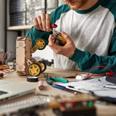 Kırpılmış genç erkek bilişim teknisyeni veya mühendis masadaki tekerlek robotunun bir parçasını dizüstü bilgisayar, çeşitli teknik aletler ve parçalarla onarıyor. Modern teknoloji ve yenilik