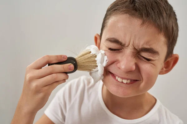 Cropped of smiling caucasian little boy smearing shaving foam on face with brush. Male child of zoomer generation. Modern youngster lifestyle. Skin care. White background. Studio shoot. Copy space