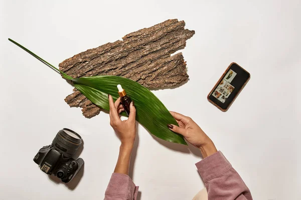 Cropped girl photographer setting up oil bottle, tree bark and leaf before taking picture on camera or smartphone at home studio. Beauty and body care content for photostocks, commerce and advertising