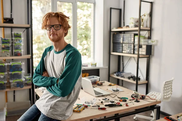 Male IT technician or engineer with crossed arms looking at camera in office. Young red haired caucasian man wearing glasses standing by table. Break and rest on work. Modern technology and innovation