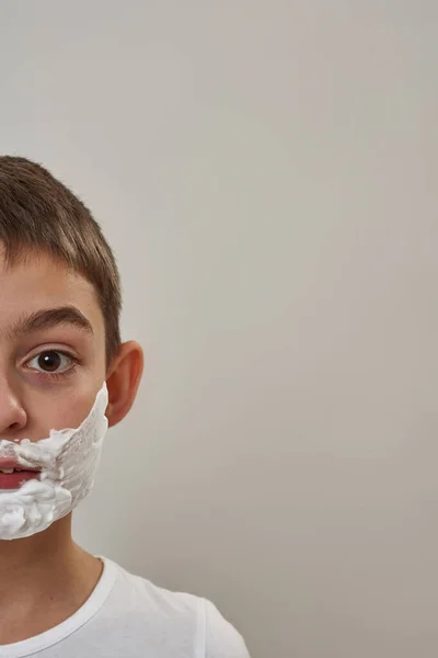 Obscure face of handsome caucasian little boy with shaving foam on face looking at camera Male child of zoomer generation. Modern youngster lifestyle. Skin care. White background in studio. Copy space