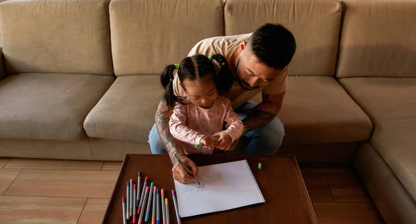 Dad and daughter draw with felt-tip pens in copybook at table at home. Asian man and little girl spend time together. Domestic entertainment and leisure. Family relationship. Parenting and fatherhood