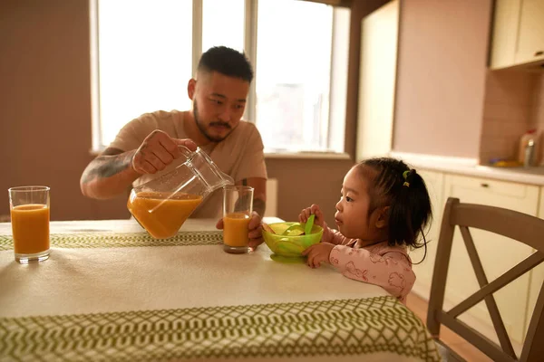 Asian father pouring juice from jar in cup while her little daughter eating cheese curd fritter on breakfast at table on home kitchen. Family relationship. Parenting and fatherhood. Healthy eating