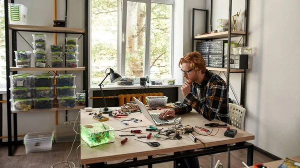 Male IT engineer typing on laptop in office. Young red haired caucasian man wearing glasses sitting at table with variety technical tools and components. Modern technology and innovation