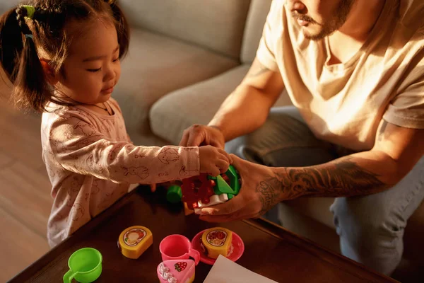 Partial father and asian daughter play with toy cube with figures at table at home. Man and girl spend time together. Domestic entertainment and leisure. Family relationship. Parenting and fatherhood