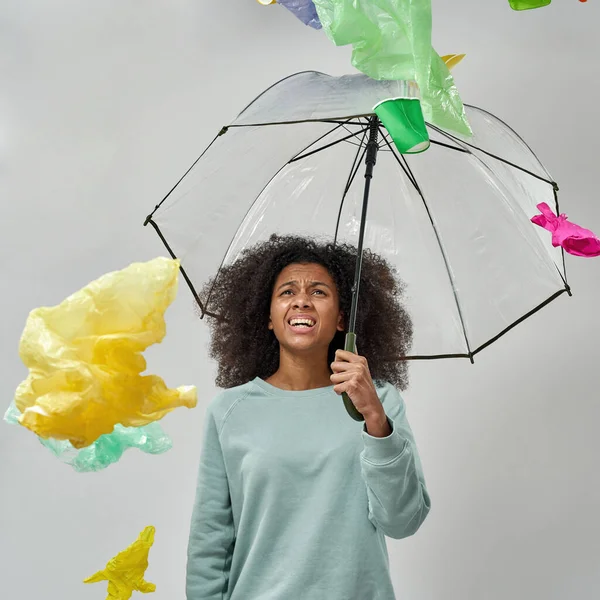 Dissatisfied black girl with umbrella standing under garbage rain. Ecology safety and protection. Waste disposal and recycling. Environmental sustainability. Isolated on white background. Studio shoot