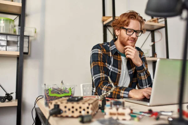 Male IT technician using and watching laptop in office. Young red haired caucasian man wearing glasses sitting at table with variety technical tools and components. Modern technology and innovation