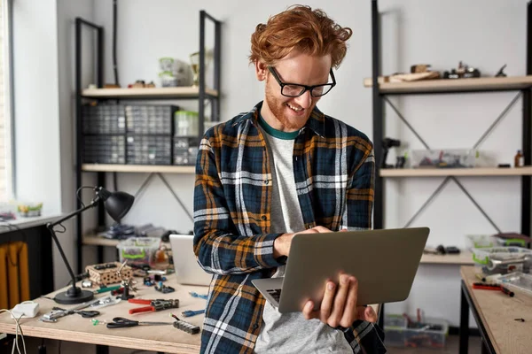 Smiling male IT engineer using laptop in office. Young red haired caucasian man wearing glasses standing by table with variety technical tools and components. Modern technology and innovation