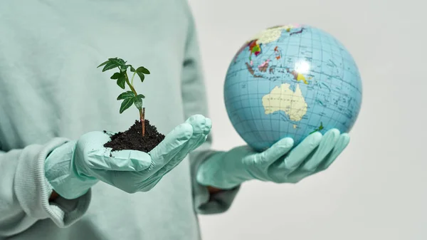 Cropped black girl wearing gloves holding earth globe and green plant in ground. Global ecology safety. Waste disposal and recycling. Environmental sustainability. White background. Studio shoot