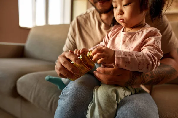Girl take multivitamin jelly from jar in cropped father hands while sit in dad arms on sofa at home. Asian man and little girl. Family relationship. Health and medical care. Parenting and fatherhood