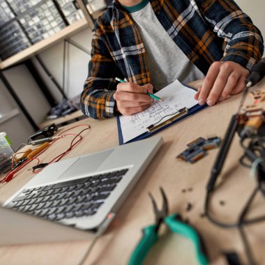Kesilmiş erkek bilişim teknisyeni kurşun kalemle teknik düzenleme yapıyor. Masada dizüstü bilgisayarla, çeşitli teknik aletlerle ve ekipmanlarla çalışan bir adam. Modern teknoloji ve yenilik gelişimi