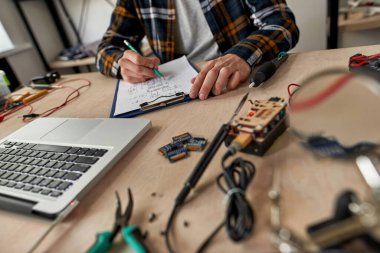 Kısmi erkek bilişim teknisyeni veya mühendis kalem ile teknik düzenleme yapıyor. Masada dizüstü bilgisayarla, çeşitli teknik aletlerle ve ekipmanlarla çalışan bir adam. Modern teknoloji ve yenilik