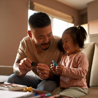 Baba, evdeki masada plastikle heykel yaparken kızına bakıyor. Asyalı bir adam ve küçük bir kız birlikte vakit geçiriyorlar. Ev eğlencesi ve eğlence. Aile ilişkisi. Babalık