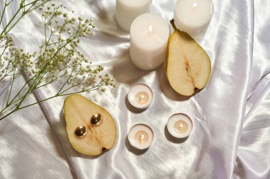 Beyaz ipek arka planda izole edilmiş armut, altın toplar ve çiçek bitkileriyle yanan mumların üst görüntüsü. Fotostoklar, ticaret, sosyal ağlar ve reklamlar için zarif bir fotoğraf. Stüdyo çekimi