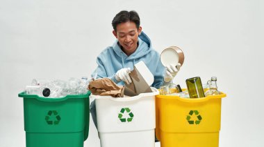 Asyalı adam çöpü cam, plastik ve karton çöp kutularına ayırıyor. Ekoloji güvenliği. Çöp öğütücü ve geri dönüşüm. Çevresel sürdürülebilirlik. Beyaz arka planda izole edilmiş. Stüdyo çekimi. Boşluğu kopyala