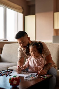 Baba-kız, evdeki masadaki kanepeye keçeli kalemlerle keçeli kalem çiziyor. Asyalı bir adam ve küçük bir kız. Ev eğlencesi ve eğlence. Aile ilişkisi. Ebeveynlik ve babalık