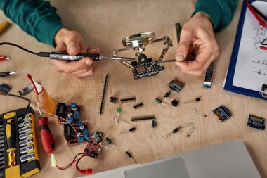 Top view of partial male technician or engineer soldering microchip with soldering iron under magnifying glass at table with laptop, variety technical tools and components. Technology and innovation clipart