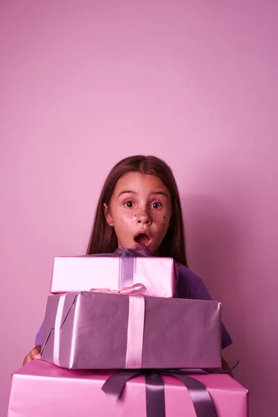 Shocked european little girl with gift boxes looking at camera. Female child with colorful sequins on face. Holiday and event. Modern youngster lifestyle. Pink background. Studio shoot. Copy space