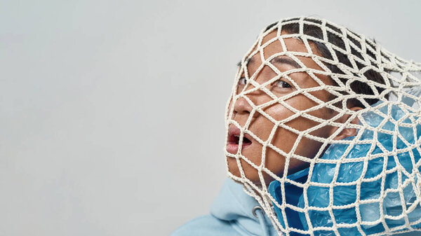 Cropped worried asian man with knitted bag full of plastic garbage on head. Ecology safety and protection. Waste disposal and recycling. Isolated on white background. Studio shoot. Copy space