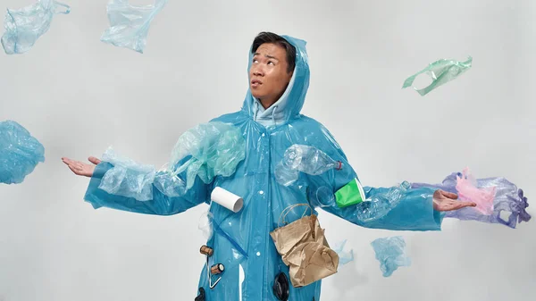 Displeased asian man wearing raincoat standing under falling garbage. Ecology safety. Waste disposal and recycling. Environmental sustainability. Isolated on white background. Studio shoot