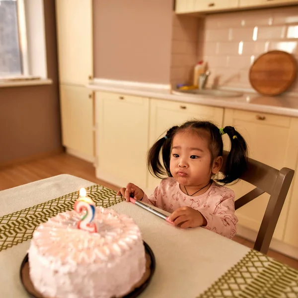 Lonely sad little asian girl sitting and looking at birthday cake with burning number candle. Upset female kid celebrating her two years old alone. Holiday and event. Modern kitchen in spacious flat